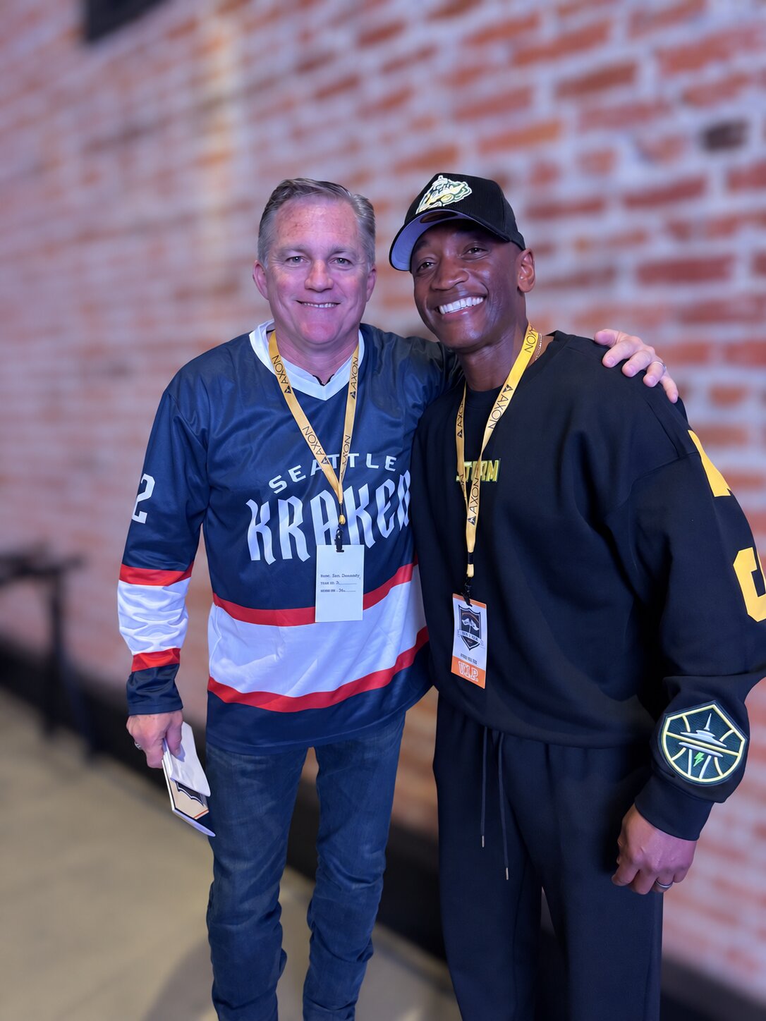 Two men smiling together at an event, one in a Seattle Kraken jersey and one in a Storm cap